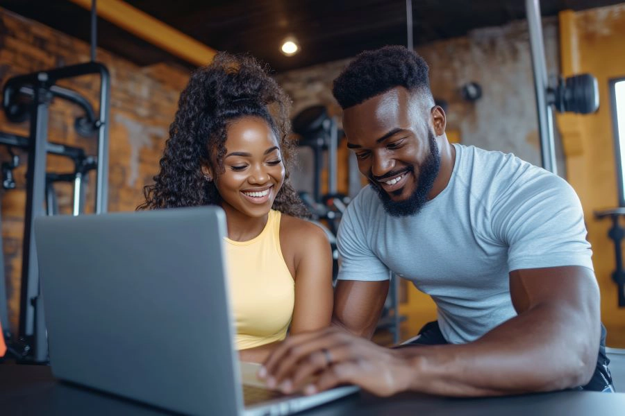 Two fit young people in a gym, with the man operating a laptop while the woman smiles,